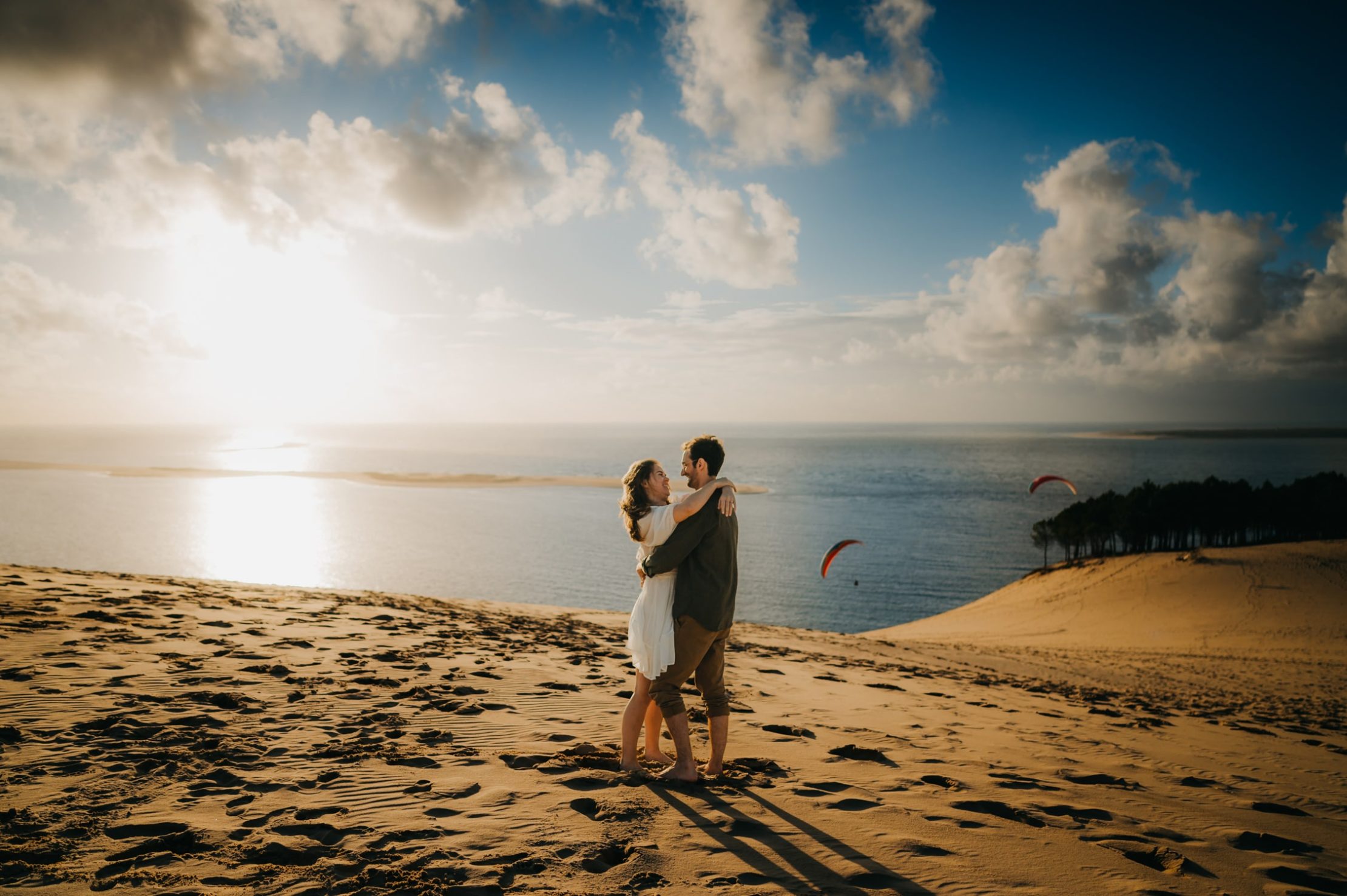 Une photographe couple à Arcachon qui fige un couple à la dune du pilat, avec des parapentes en arrière plan, lumières dorés et ambiance romantique