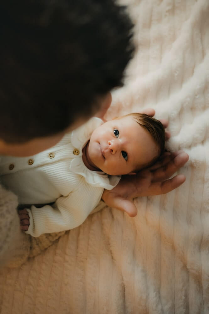 Un papa tient son bébé dans les bras. Son bébé le regarde dans les yeux. Un instant tendre capturé pendant une séance photo naissance par L'instant par Phé, photographe bébé Bordeaux et Gironde.