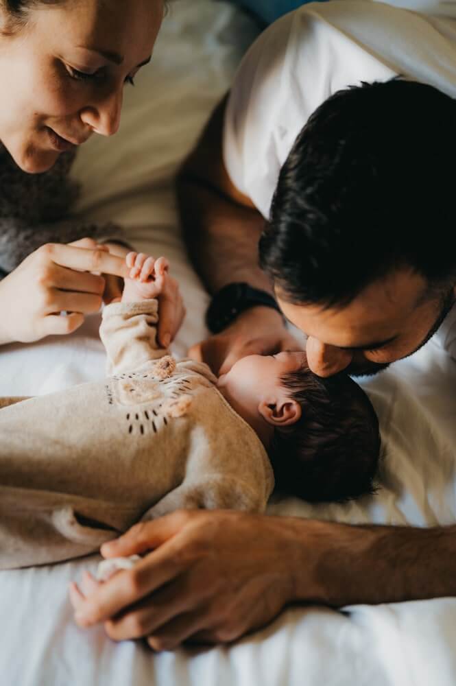 Les parents sont sur le lit avec leur bébé pendant leur séance photo naissance. Le papa embrasse son bébé sur le front. Capturé par L'instant par Phé, photographe bébé Bordeaux et Gironde.