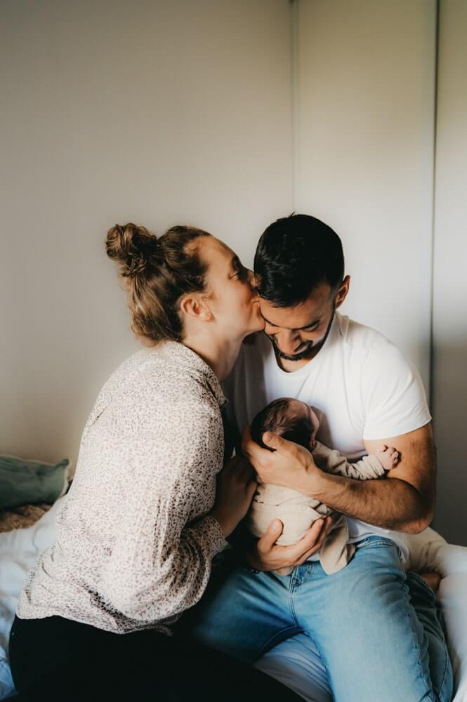 Le papa tient son bébé contre lui pendant que la maman l'embrasse sur la joue. Une séance photo naissance par L'instant par Phé, photographe bébé Bordeaux et Gironde.